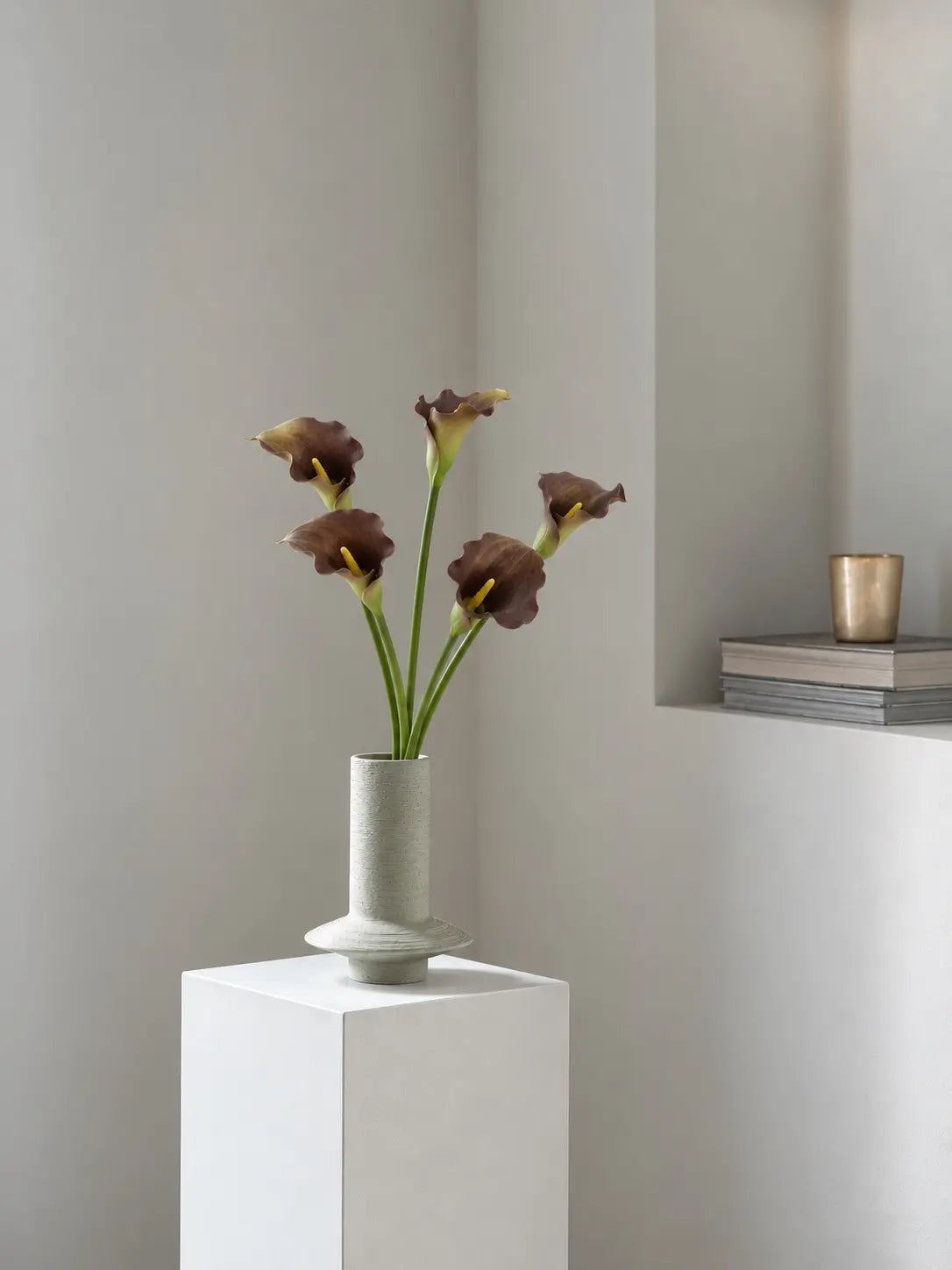 Five brown faux calla lilies arranged in a sculptural pale vase on a white plinth