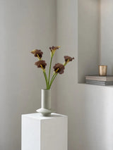 Five brown faux calla lilies arranged in a sculptural pale vase on a white plinth